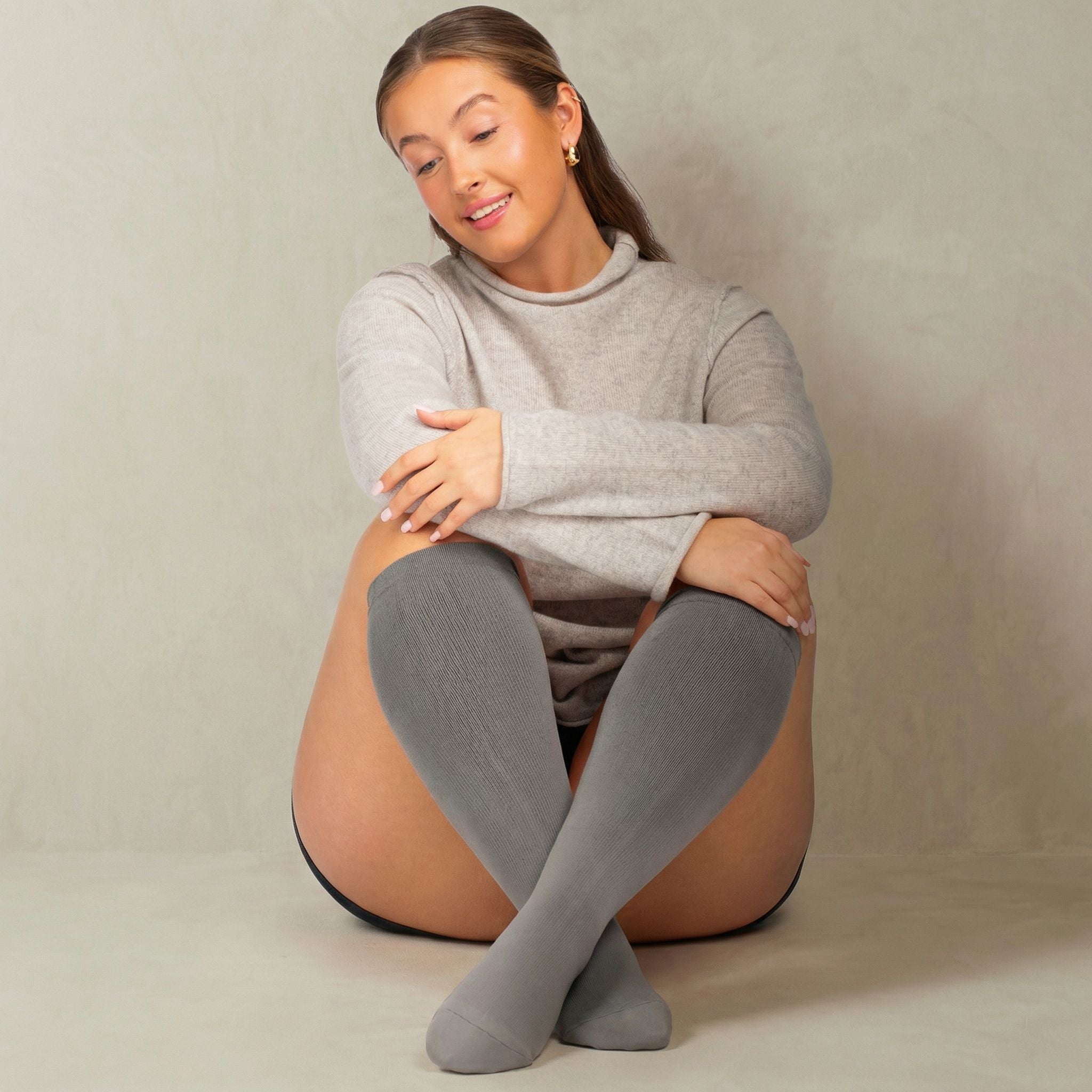 Woman wearing gray compression socks sitting on a beige surface with a neutral background