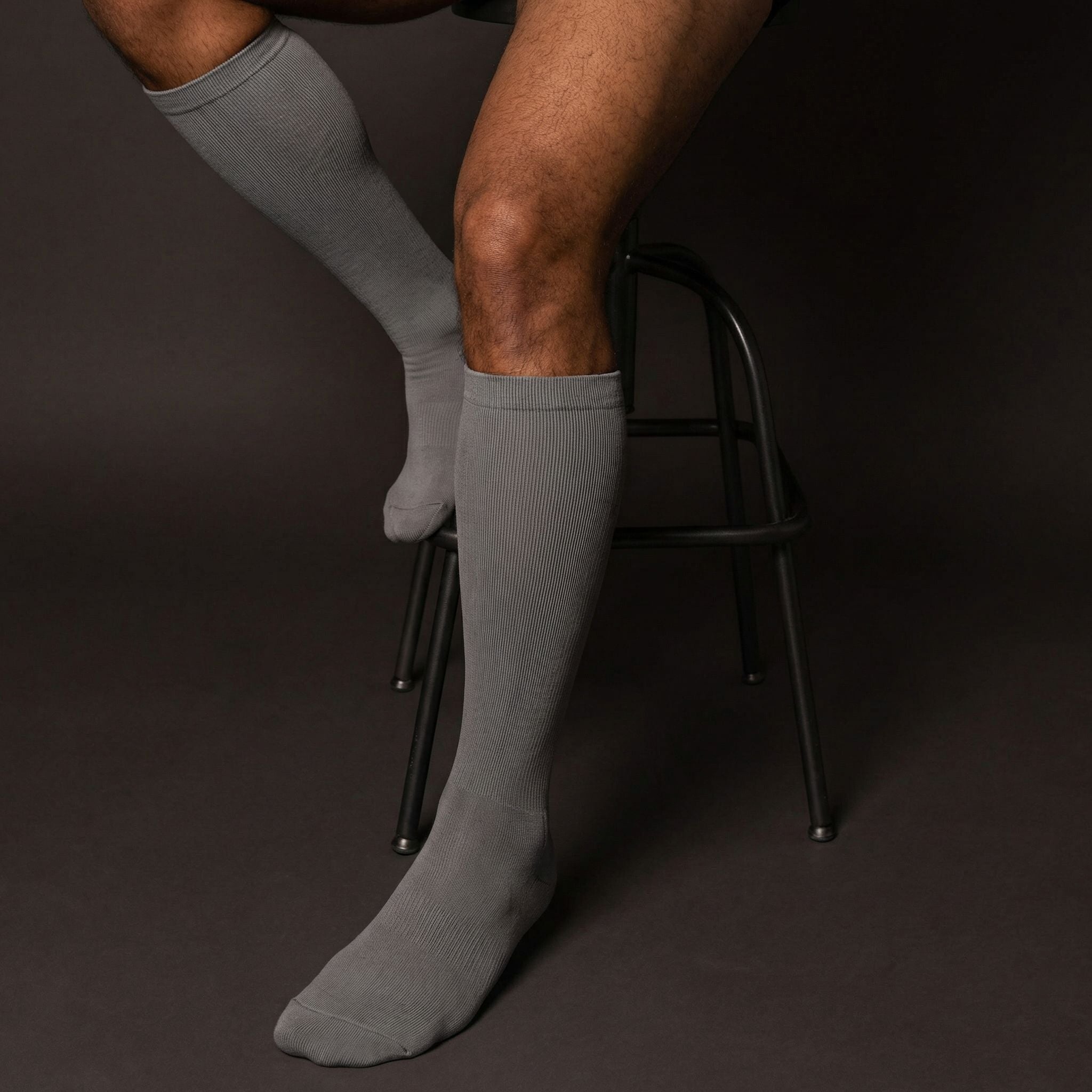 Person wearing gray compression knee-high socks sitting on a black stool against a dark background