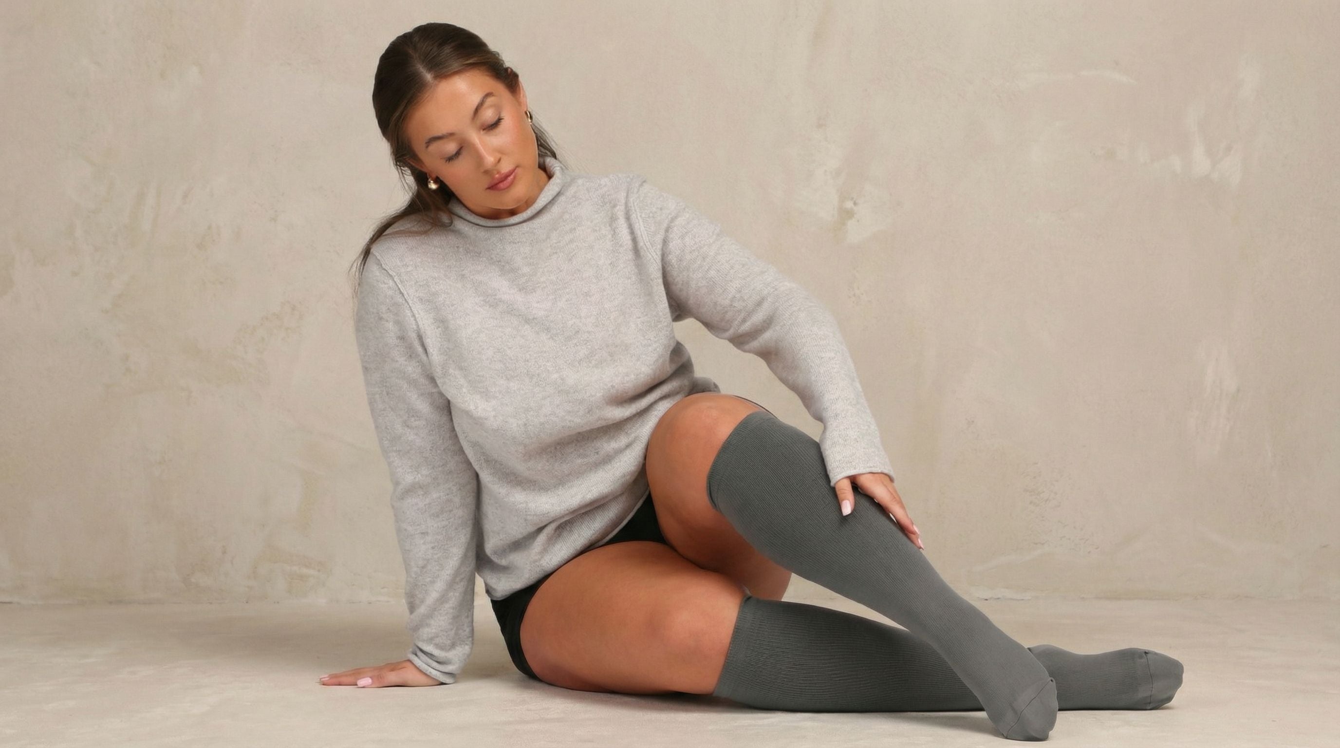 Woman wearing a gray sweater and knee-high compression socks sitting on the floor against a plain background